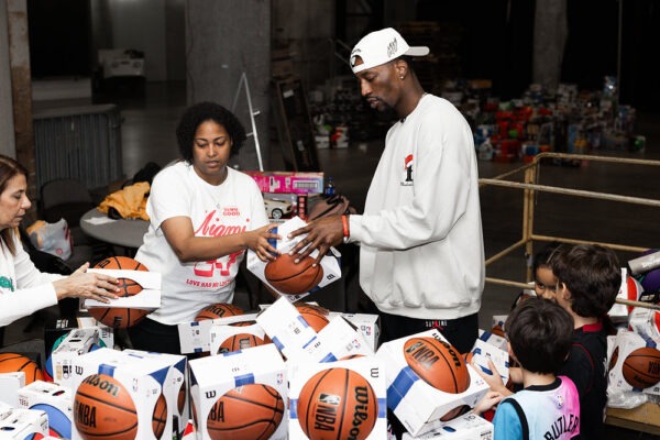 Bam Adebayo stands on the court at Kaseya Center surrounded by children and tables filled with holiday toys during a large community giveaway.